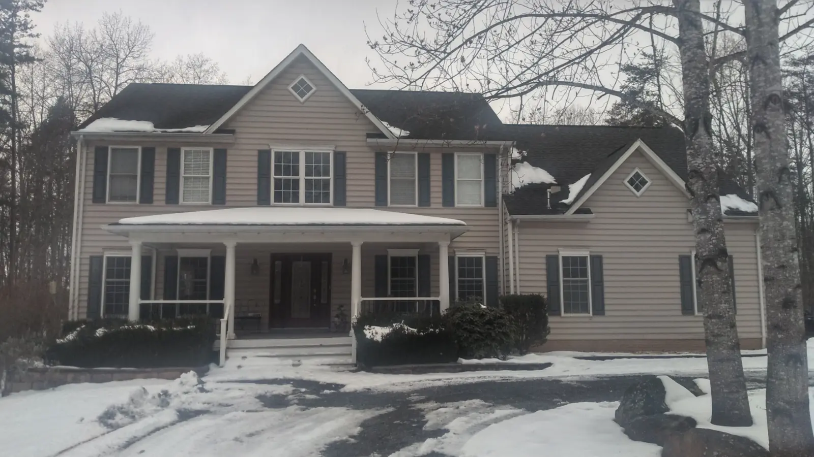 Wide view of a two-story beige home with a covered porch, dark shingle roof and snow on the ground.