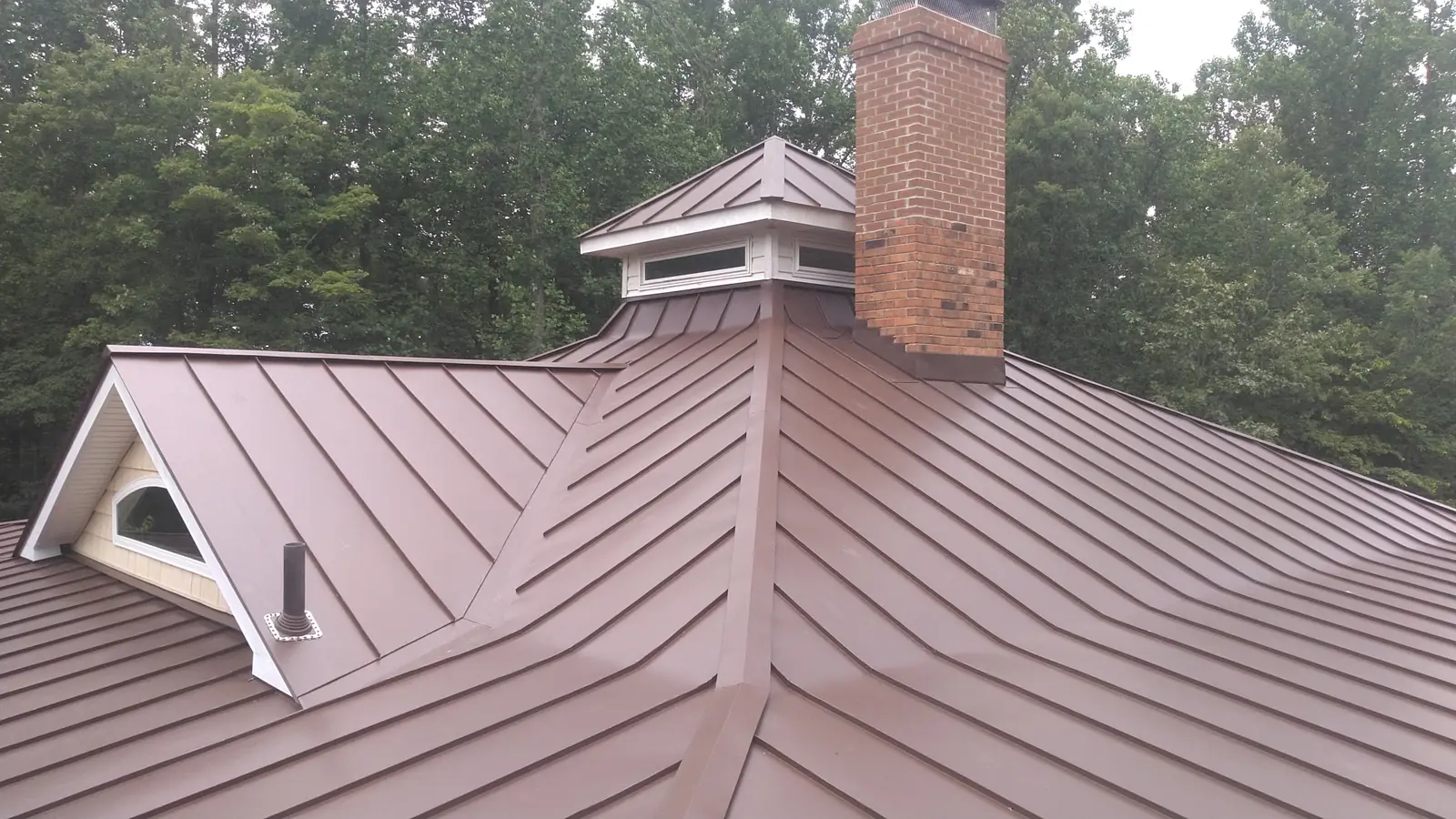 Wide view of bronze standing seam roof with an arched dormer, cupola and chimney.