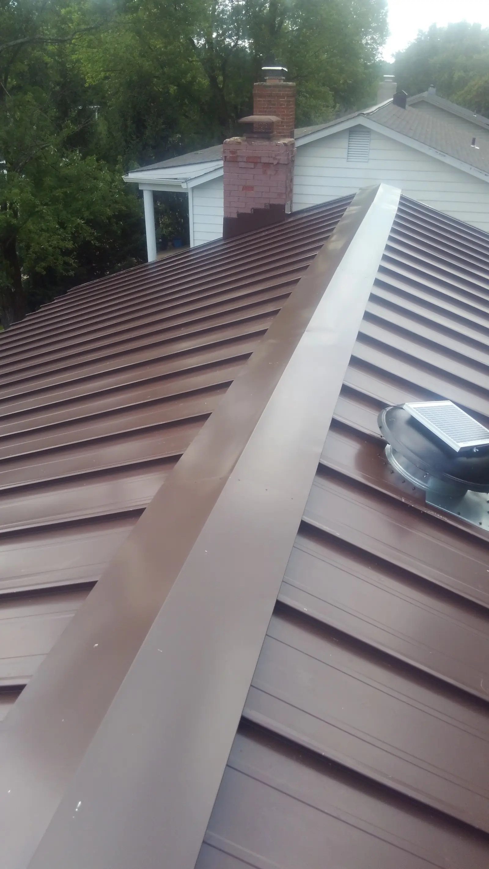 Brown standing seam ridge with a vent and a chimney in the distance.
