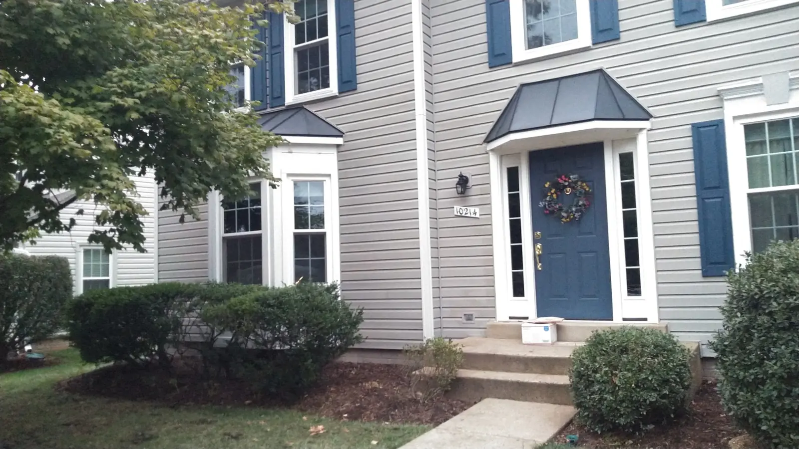 Gray-sided home with a blue door under a dark metal hood and a bay window beside it.