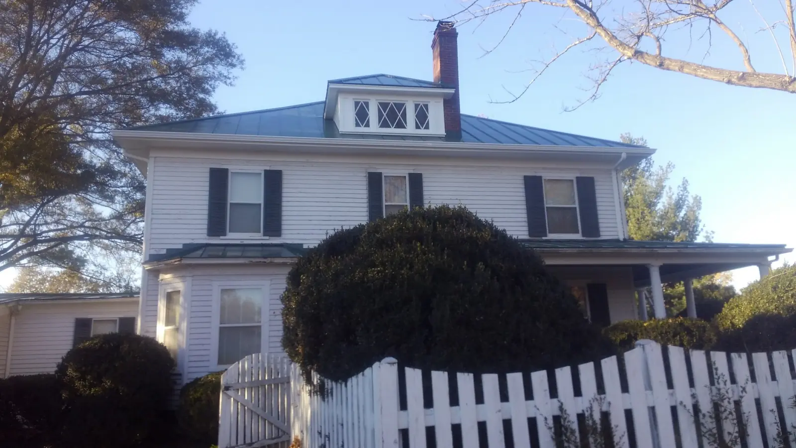 White colonial with a green metal hip roof, a porch and a picket fence.