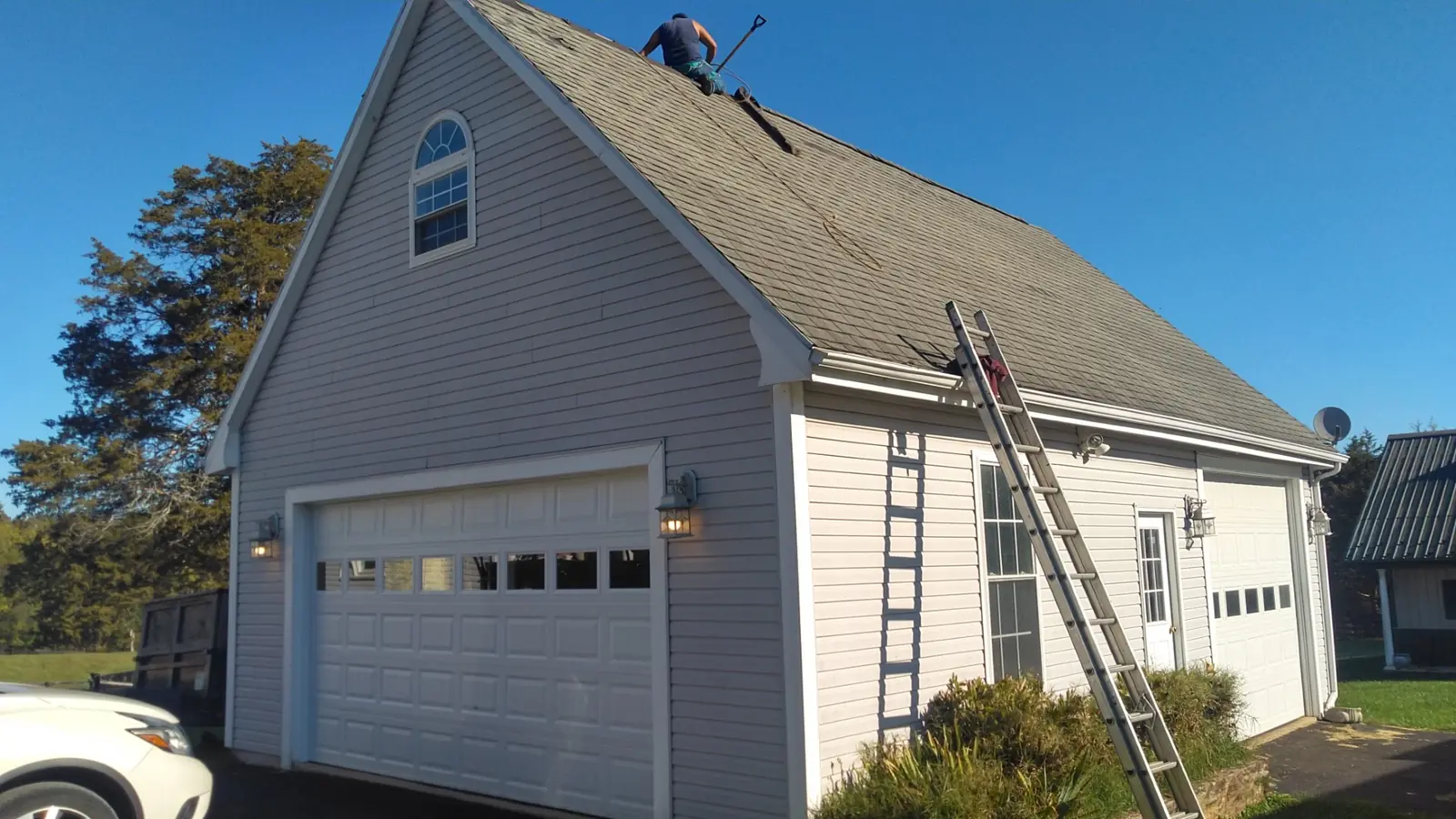 Garage before the work with a gray shingle roof, a ladder and a crew member on top.