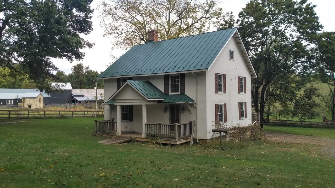 Farmhouse with a green metal roof, porch roof and trees behind.