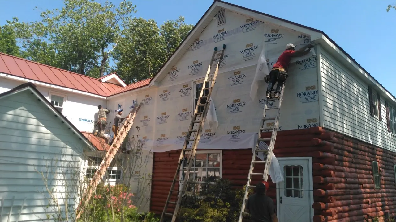 Crew on two ladders wrapping the gable end of a house with red metal roofs.