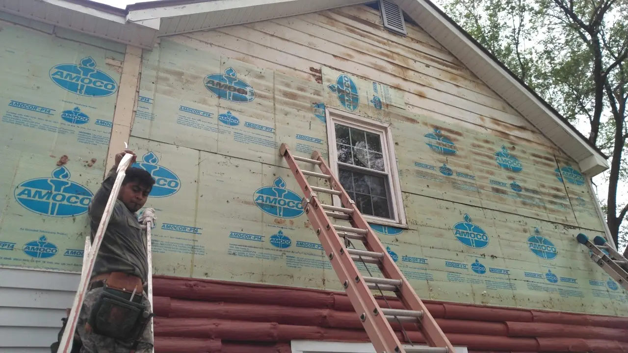 Worker on a ladder beside house wrap over a wall with red log-look siding below.