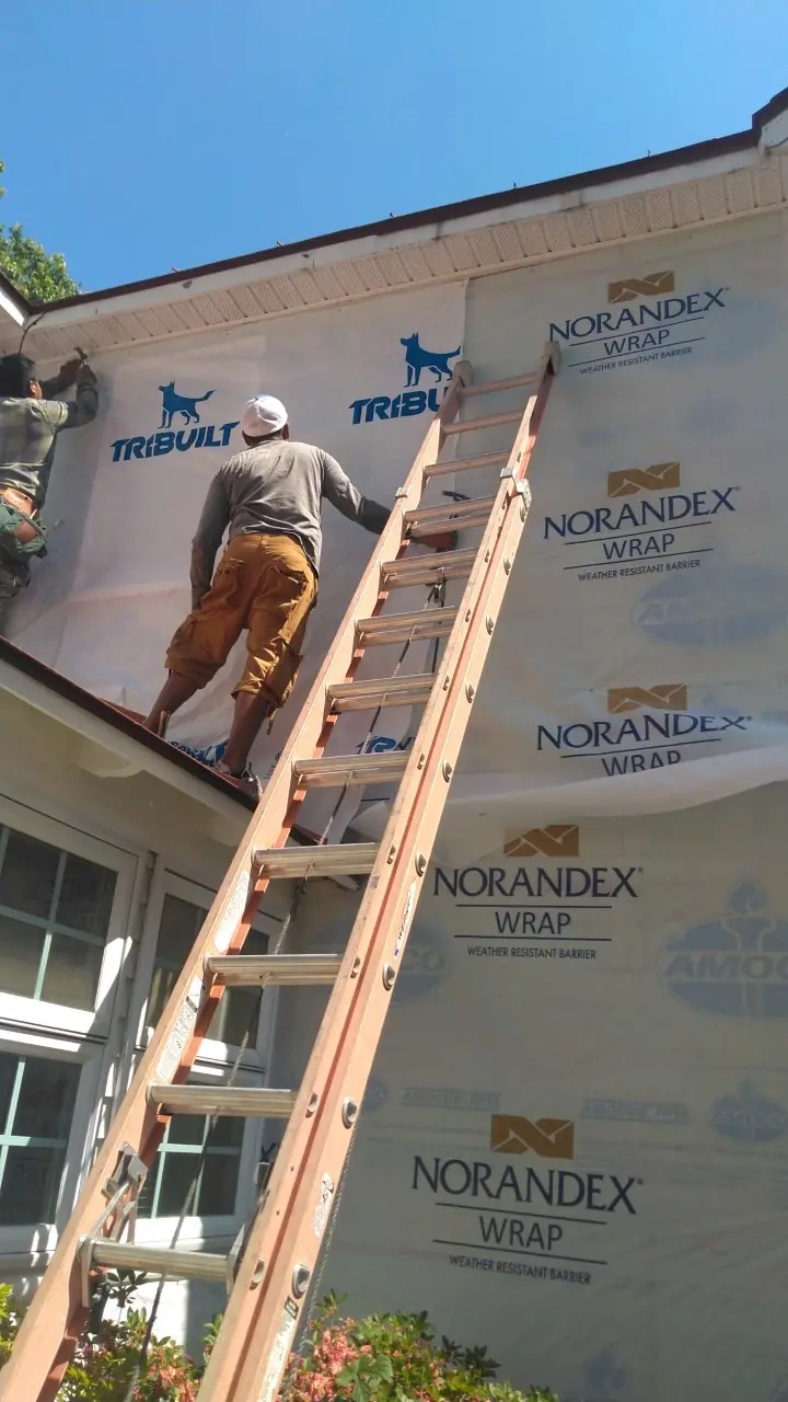 Crew member on a ladder fastening house wrap on a second-story wall.