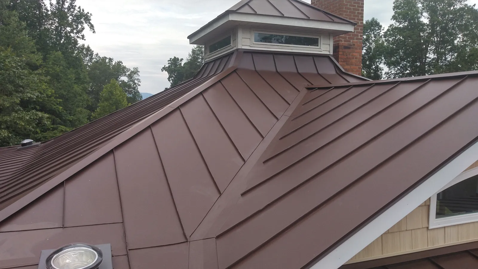 Brown standing seam hip roof with a skylight dome and chimney in the background.