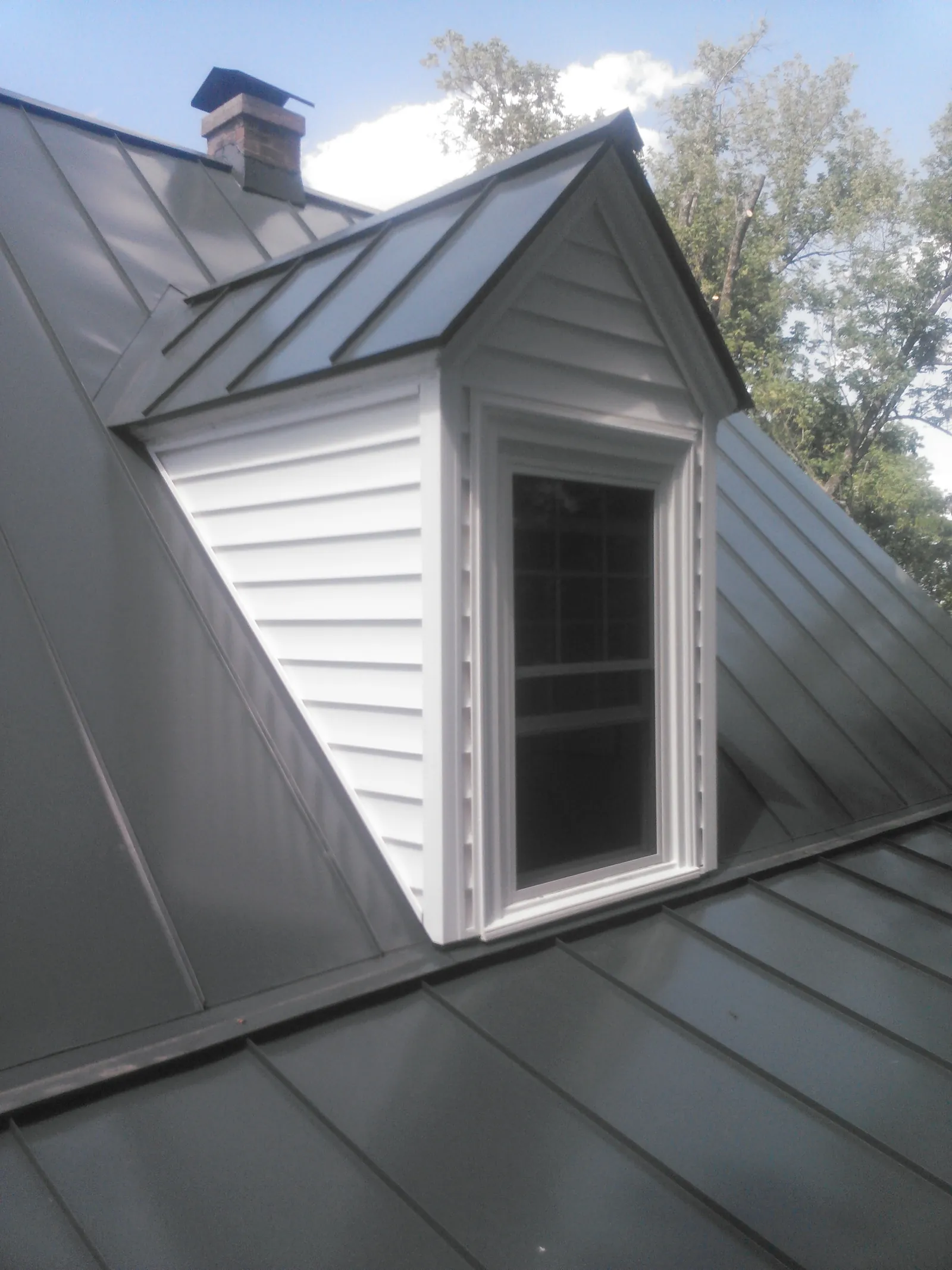 Dark standing seam roof around a white vinyl-sided dormer with a window.