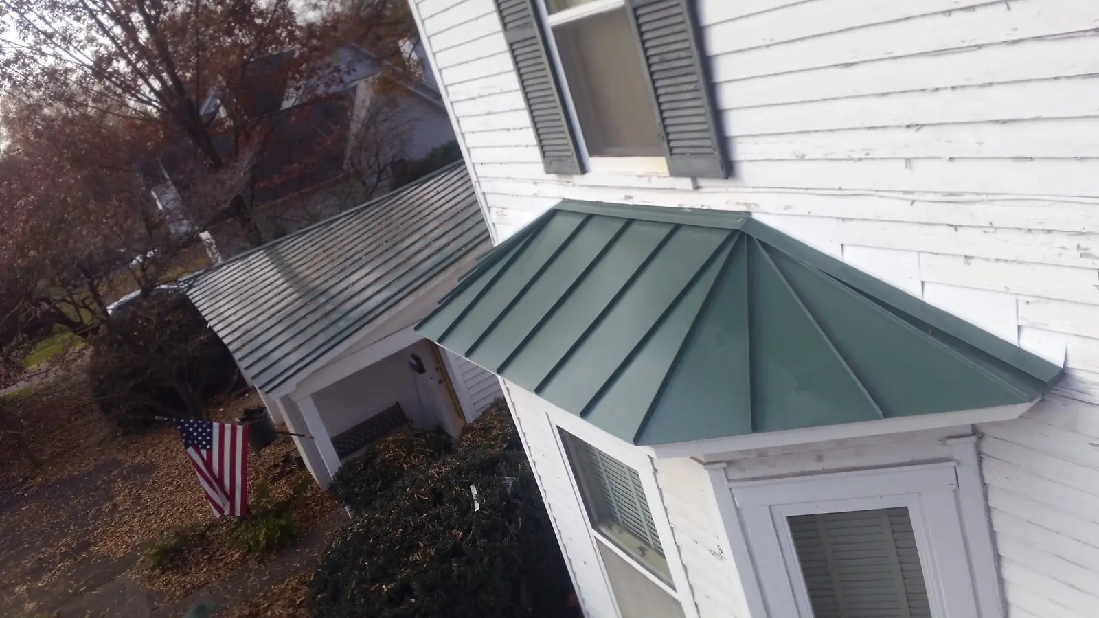 Green standing seam roof over a bay window beside a porch roof on a white house.