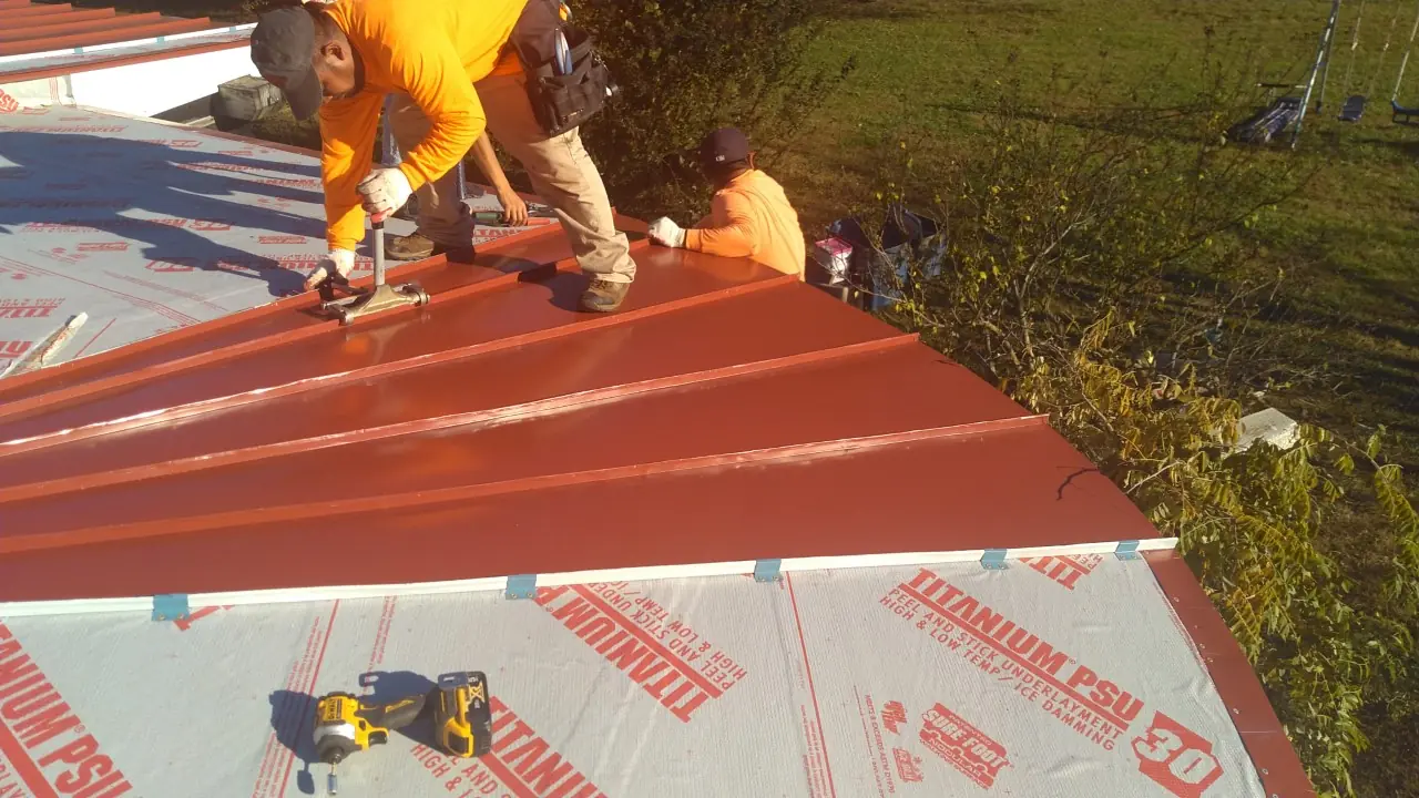 Crew installing red standing seam panels over underlayment with a drill and fasteners.