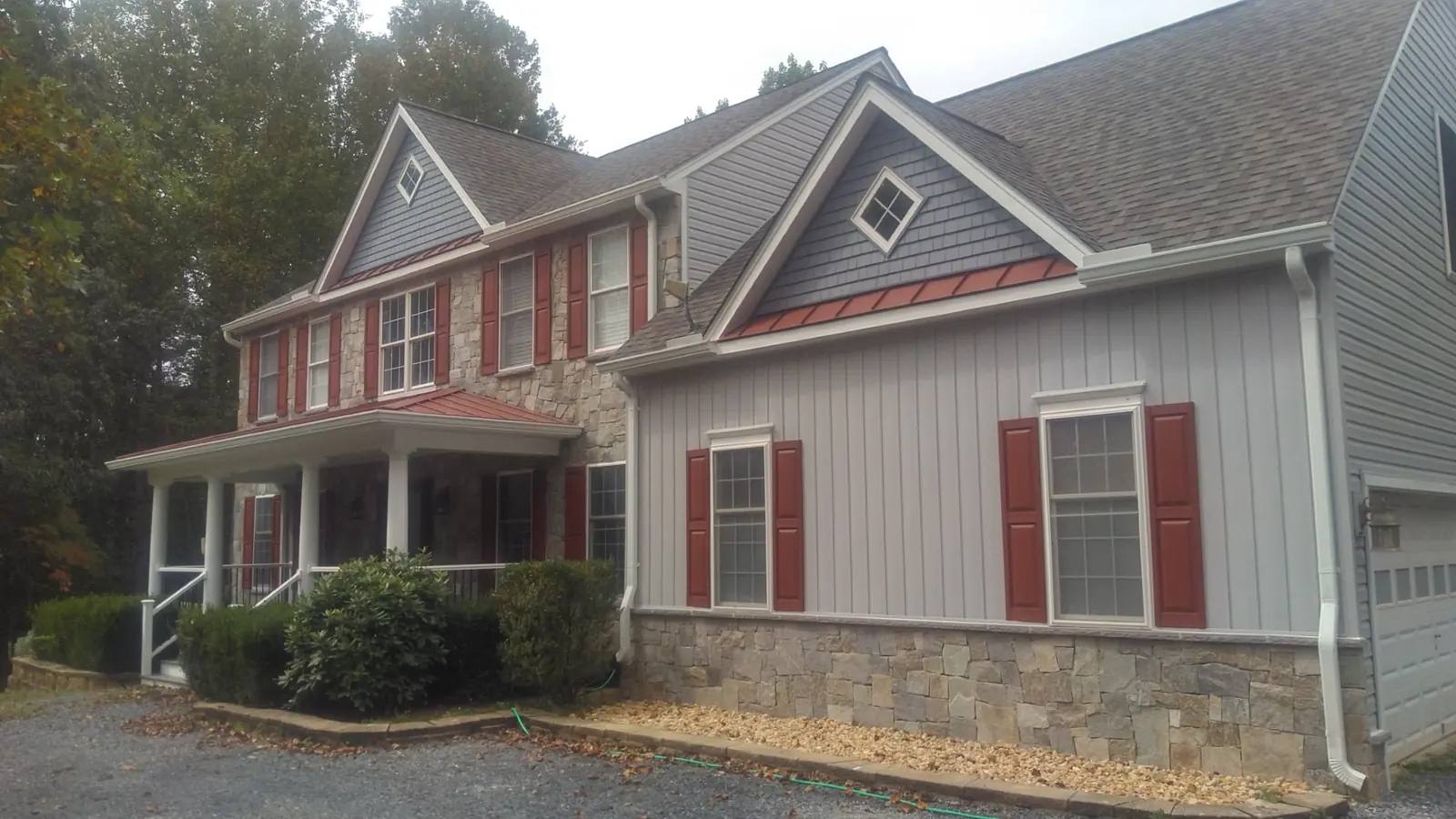 Gray board-and-batten home with a stone base, red shutters and a red metal accent roof over a gable window.