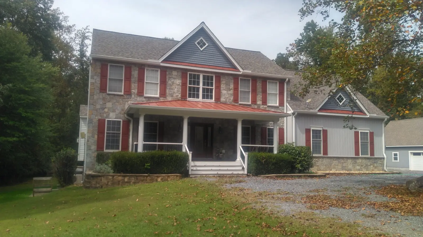 Home with a red metal porch roof and gable, stone foundation and red shutters seen across a gravel drive.