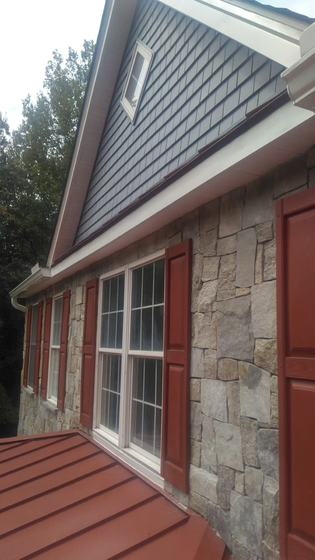Dark blue-gray gable siding above stone and a red standing seam roof.
