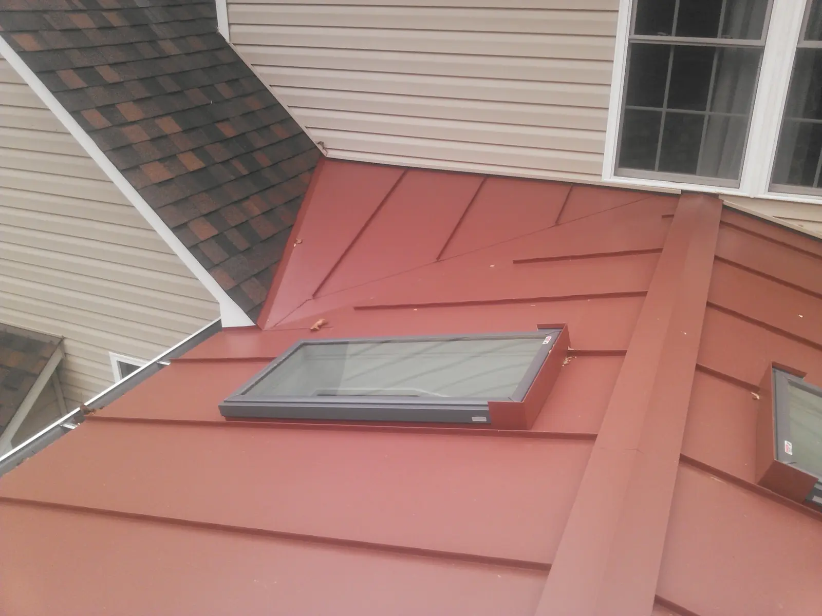 Red standing seam roof with a skylight beside a brown shingle roof.