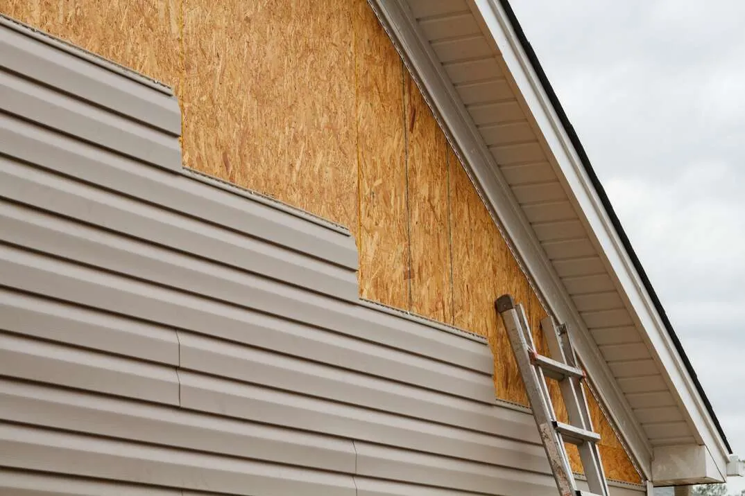 Vinyl siding removed on a gable end showing sheathing during a repair, with a ladder alongside.