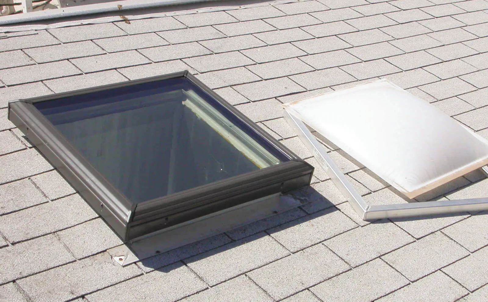Framed skylight beside its removed dome on a light grey shingle roof.