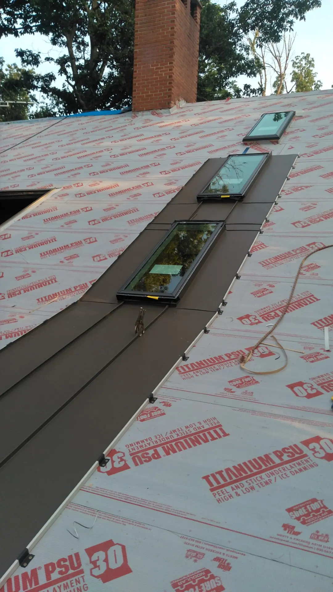 Row of skylights set in brown standing seam panels on underlayment beside a brick chimney.