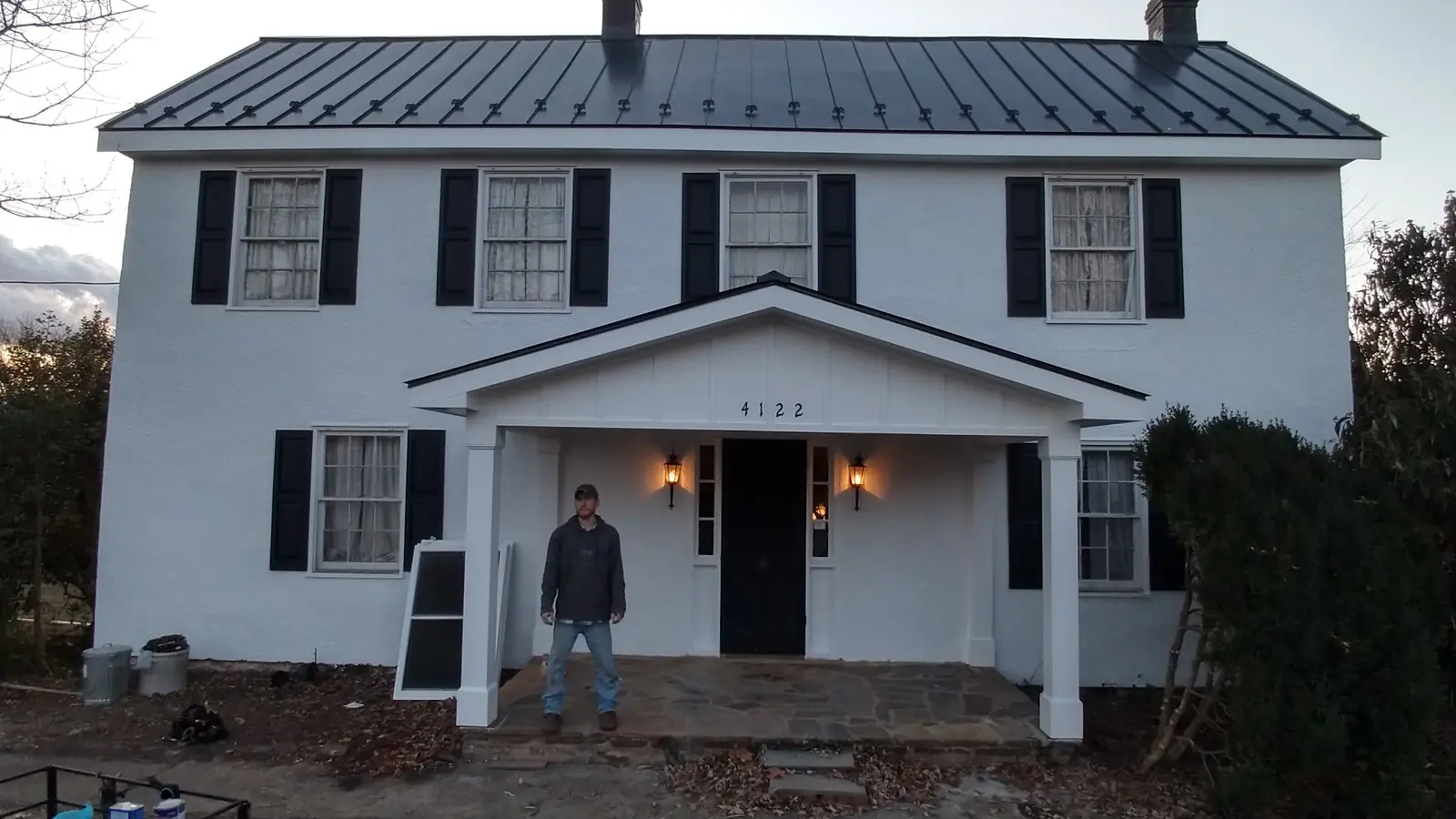 Front porch of a white colonial with a dark metal roof; a person stands at the door.