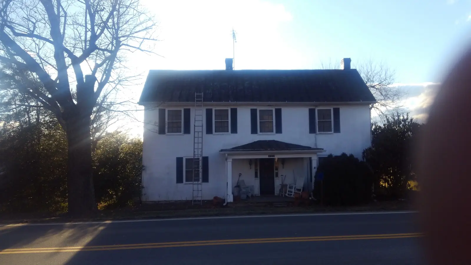 White colonial seen from the road with a ladder at the porch.