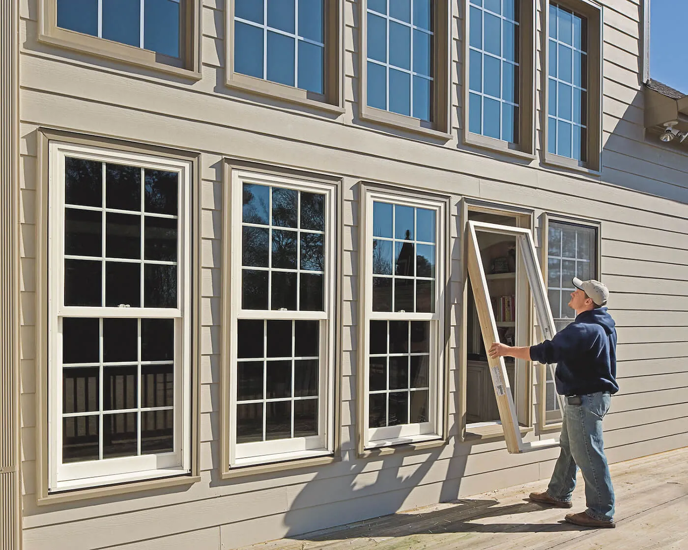 Tall double-hung windows on tan siding while a worker handles a window sash on the deck.